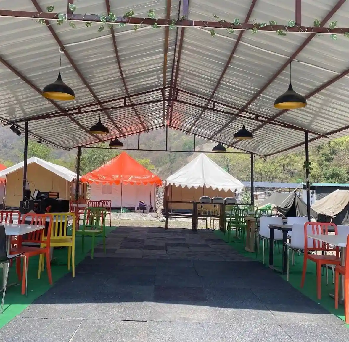 overed open-air dining area at CampNStay with colorful chairs and tables, hanging lights, and luxury tents set against green hills in the background.