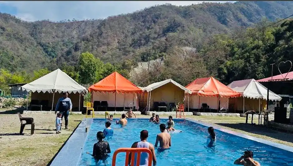 Guests enjoying a swimming pool at CampNStay riverside campsite, with luxury tents and forest-covered hills in the background.