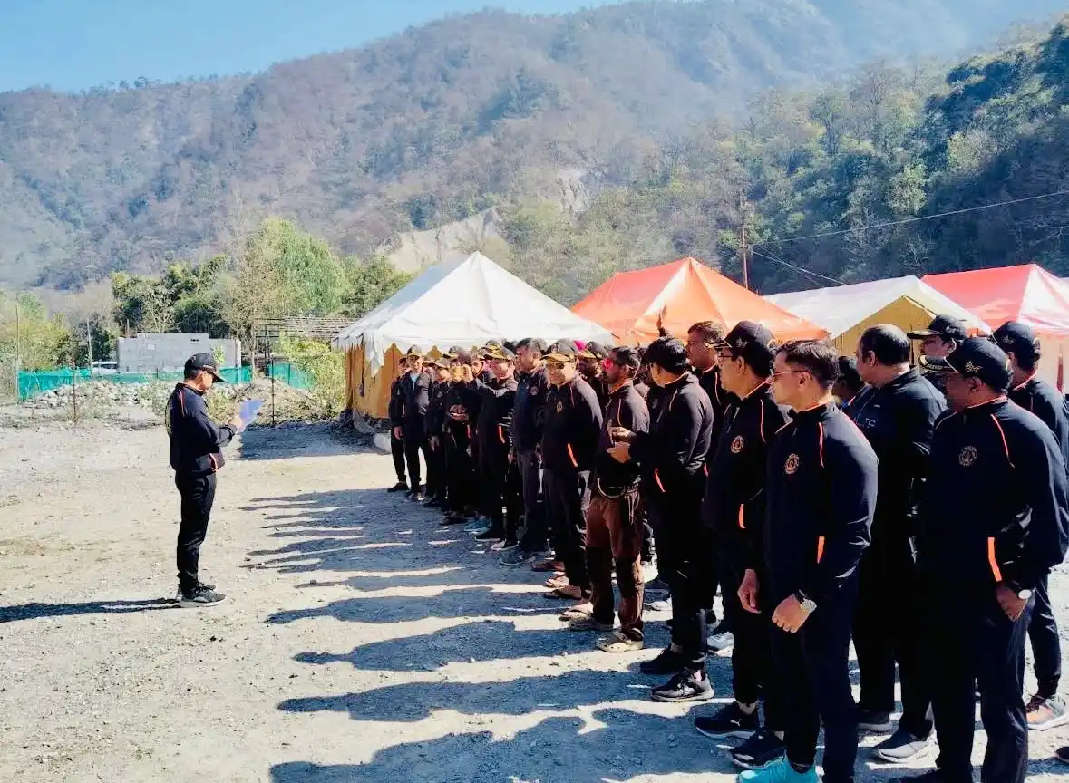 Group of uniformed campers standing in a line during a briefing at a CampNStay riverside campsite, with tents and forested hills in the background.